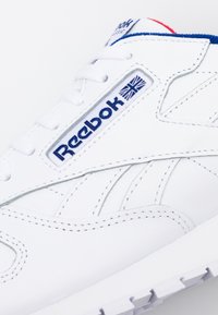 White Reebok Classic sneaker with blue and red accents, visible laces, and Reebok logo with Union Jack flag on the side.