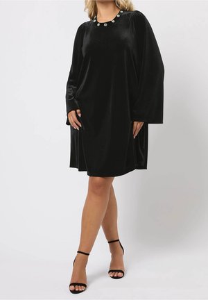 Woman wearing a black velvet long-sleeve dress with floral neckline details and black high-heeled ankle-strap sandals, standing.