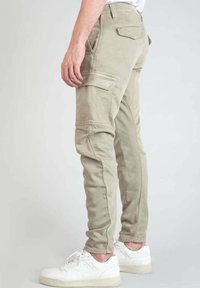 Pantalones cargo color caqui con múltiples bolsillos, diseño ajustado, textura sin costuras y sutilezas en los detalles de la costura, combinados con zapatillas deportivas blancas.