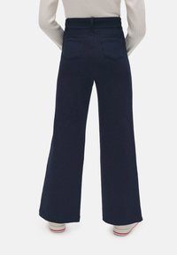 Pantalones acampanados de color azul marino, confeccionados en una tela suave y con una cintura alta, que presentan dos bolsillos traseros y un diseño de pernera ancha, combinados con zapatos de color claro.