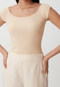 Vaahto beige -pusero, jossa on lyhyet hihat ja neliömäinen pääntie, yhdistettynä vaaleanvärisiin housuihin, joissa on sileä pinta ja joustava vyötärö.