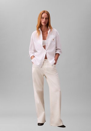 Camicia bianca con bottoni e maniche arrotolate, abbinata a pantaloni ampi color crema e scarpe scure. Dettaglio logo dorato sulla tasca.