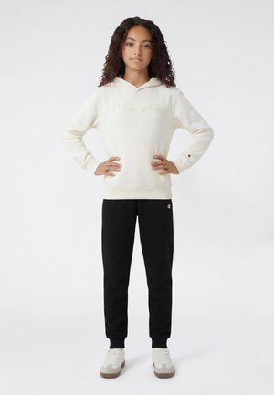 Junge Person steht mit den Händen in den Hüften, trägt einen cremefarbenen Hoodie, schwarze Jogginghosen und weiße Sneaker vor einem schlichten weißen Hintergrund.