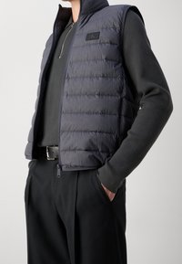 Ciemnoszara puchowa kamizelka z wysokim kołnierzem, zapinana na zamek błyskawiczny, z naszywką z logo CK, noszona na czarnej koszuli z długim rękawem i czarnych spodniach.