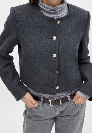 Persona que lleva una chaqueta abotonada texturizada de color gris oscuro sobre un jersey de cuello alto gris claro, con pantalones grises y un cinturón marrón, manos en los bolsillos.