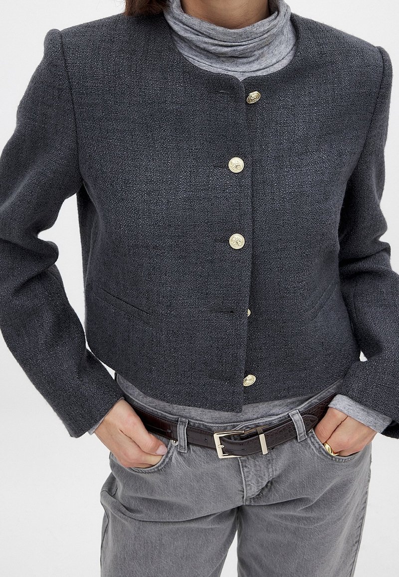 Personne portant une veste boutonnée texturée gris foncé sur un col roulé gris clair, avec un pantalon gris et une ceinture marron, mains dans les poches.