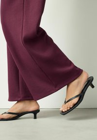 Čierne ploché sandále s tenkými remienkami a nízkym podpätkom, v kombinácii so širokými bordovými nohavicami. Viditeľné sú hladké textúry látky.