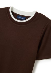 T-shirt marrone a maniche corte con un colletto a girocollo bianco a costine; realizzato in un morbido tessuto. Presenta un'etichetta cucita con "PIOMBO" in blu.