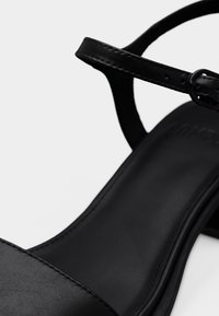 Μαύρο δερμάτινο πέδιλο με ψηλό τακούνι, λουράκι στον αστράγαλο και αγκράφα, με λεία επιφάνεια πέλματος και λεπτομερείς ραφές.