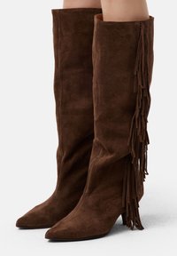 Botas de ante marrón hasta la rodilla con puntera afilada y un diseño acampanado, que presentan detalles de flecos laterales y un tacón bajo.