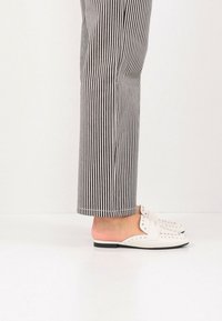 Chaussures blanches à enfiler avec des accents cloutés, présentant une finition lisse et une semelle basse noire. Portées avec un pantalon rayé noir et blanc.