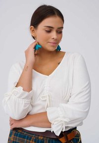 Witte blouse met lange mouwen, voorzien van geplooide details en koordaccenten bij de taille en manchetten. Teal-kleurige bloemenoorbellen. Ruitpatronen rok zichtbaar.
