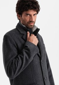 Abrigo tricolor gris oscuro con un diseño estructurado, que presenta un cuello alto y cierres de botones. La tela tiene una textura suave con una capa interior de contraste.