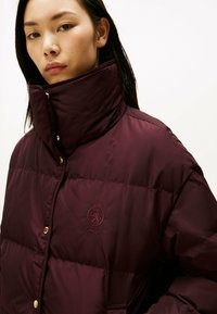 Kobieta ubrana w burgundową kurtkę puchową z wysokim kołnierzem i złotymi zatrzaskami, z ukośnym haftem herbu z lwem oraz napisem na piersi.