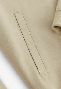 Gros plan d’un tissu beige avec une poche passepoil verticale et une partie d’un revers roulé visible sur le côté droit.