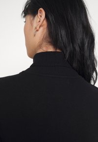 Maglione nero con collo alto, caratterizzato da una texture a coste e un design aderente. Il colletto presenta una finitura a coste, completata da capelli scuri e lucidi.
