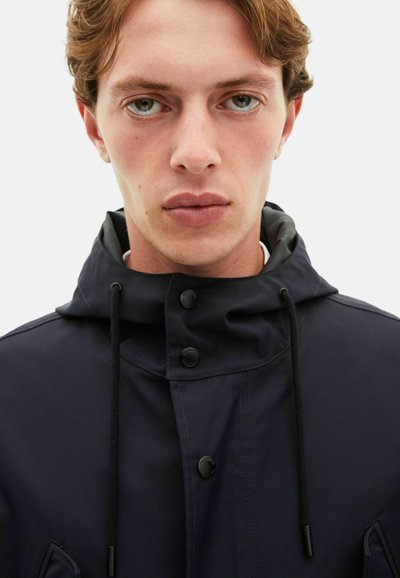 Veste imperméable bleu marine avec un col haut, un cordon de serrage réglable, des boutons-pression et une texture lisse. Conçue pour la fonctionnalité.