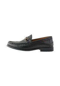 Mocasín de cuero negro con un acabado texturizado, forma alargada y un acento metálico decorativo en la parte superior. Suela plana con un mínimo relieve.