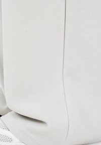 Κοντινό πλάνο λευκού υφάσματος με ρίγες, με ραφή και μέρος ενός λευκού αθλητικού παπουτσιού από δίχτυ στο κάτω μέρος.