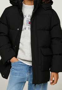 Osoba oblečená v černé péřové bundě přes šedou mikinu s modrým logem a světle modrými džíny, jedna ruka v kapse a druhá uvolněná.