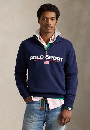 Μπλε ναυτικό φούτερ με φερμουάρ και λογότυπο "POLO SPORT" σε άσπρο και κόκκινο, φορεμένο πάνω από πράσινο και ροζ πουκάμισο με γιακά. Συνδυάζεται με ανοιχτόχρωμο τζιν.