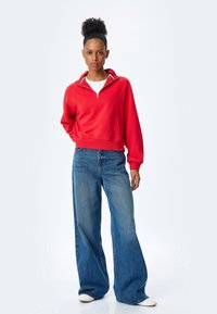 Czerwona bluzka z zamkiem błyskawicznym i wysokim kołnierzem, z ribbami na mankietach, w połączeniu z szerokimi niebieskimi jeansami i białymi trampkami.
