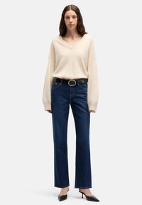 Maglione beige a scollo a V, vestibilità comoda, abbinato a jeans blu scuro a gamba dritta. Cintura nera in pelle con fibbia argentata, tacchi neri a punta.