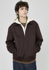 Veste zippée marron avec capuche crème, poignets et taille élastiques. Tissu lisse, poches avant et design de col classique.