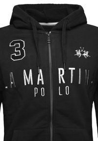 Černá zipová mikina vyrobena z měkké látky má přední kapsu, kapuci se šňůrkou a bílé vyšívané detaily včetně čísla a loga.