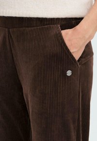 Brown corduroy pants with vertical ribbing, featuring a side pocket and a metallic logo button on the pocket. Soft texture.