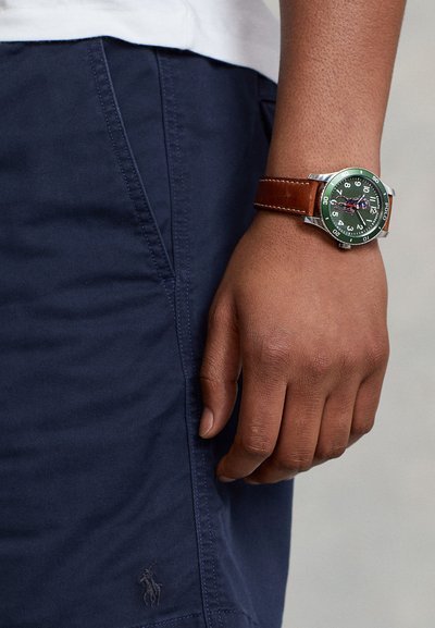 Brun klocka med läderarmband, grön urtavla, silverfärgad boett, arabiska siffror och präglad logotyp; bärs på en hand i marinblå shorts.