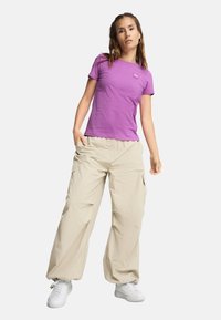 T-shirt viola a maniche corte con scollo rotondo e piccolo logo, abbinato a pantaloni cargo beige con tasche e vestibilità rilassata.