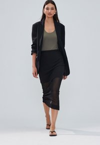 Blazer nero su una canottiera oliva e gonna a matita nera trasparente, con orlo arricciato e semplici sandali neri. Texture lisce, design minimale.