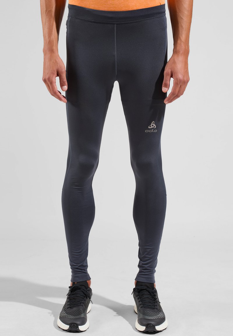 Zwarte sportleggings gemaakt van rekbare stof, met een gladde textuur en een reflecterend logo op de linker dij, gecombineerd met zwarte sneakers.