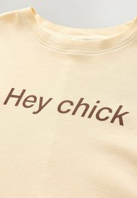 Kollane puuvillane T-särk, millel on ümar kaelus ja tumepruunis kirjas tekst "Hey chick", sile tekstuur, minimalistlik disain.