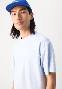 Lyseblå t-shirt med korte ærmer, der har horisontale strukturerede striber. Bærer en blå kasket. Stoffet ser blødt og afslappet ud.