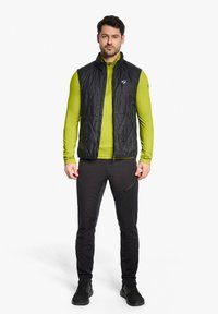 Schwarz isolierte Weste mit hohem Kragen, getragen über einem grünen Langarmshirt, kombiniert mit schwarzen Hosen und schwarzen Sportschuhen.