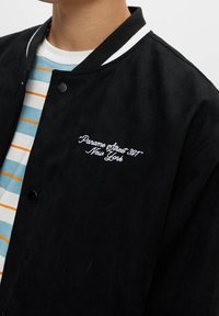 Black suede jacket with white trim and buttons; features embroidered text “Paname Street 307” and “New York.” Worn over a striped shirt.