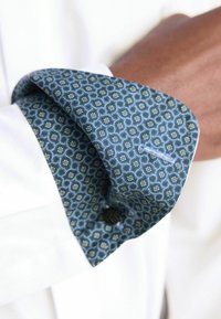 Weißer Hemdärmel mit dunkelblauer gemusterter Innenverkleidung, die florale Motive zeigt, mit Knopfdetail und strukturiertem Stoff. Der Ärmel ist umgeschlagen, um die Innenverkleidung zu zeigen.