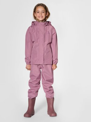 Enfant portant une veste de pluie mauve assortie, un pantalon de pluie et des bottes, debout et souriant sur un fond uni.