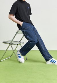 Camiseta negra, jeans azules y zapatillas blancas con rayas azules. La pose es casual, sentado en una silla plegable transparente sobre un suelo verde.