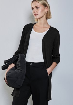 Zwarte lange cardigan van zachte stof, met een open voorzijde, gedragen over een witte tanktop, gecombineerd met zwarte broek, en met een gewatteerde tas.