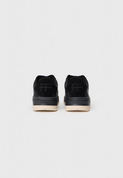Ein Paar schwarze Low-Top-Sneaker mit cremefarbenen Sohlen, von hinten auf einem schlichten hellgrauen Hintergrund gesehen.