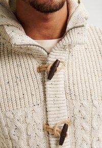 Maglione a maglia crema con un motivo a treccia testurizzata, dotato di cappuccio con cordoncino e chiusura a toggles in legno con spago.