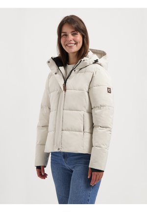 Veste blanche rembourrée avec une capuche, présentant des sections matelassées, un zip à l'avant et des accents noirs. Portée par-dessus une chemise légère et un jean.