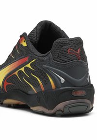 Schwarzer Sneaker aus Mesh mit roten und gelben Akzenten. Verfügt über ein geschwungenes Design, eine gepolsterte Sohle und ein strukturiertes Obermaterial mit Markenlogo an der Ferse.