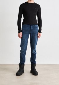 Svart långärmad tröja, blå jeans och svarta isolerade stövlar. Tröjan har en liten logotyp; jeansen har en standard passform och en liten blekning.