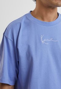 Light blue cotton t-shirt featuring a crew neckline and loose fit. It has white embroidered text on the front depicting a signature style.