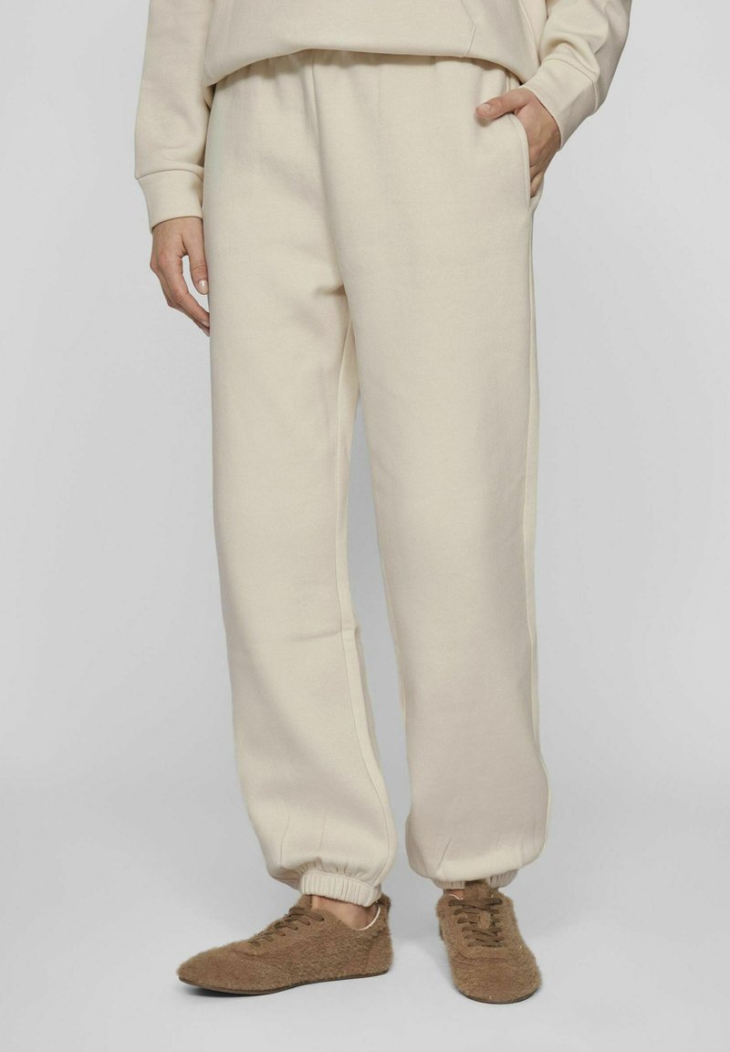 Pantalones jogger de color crema hechos de una tela suave, con puños elásticos y bolsillos laterales, combinados con zapatos de casa marrones y esponjosos que se deslizan fácilmente.