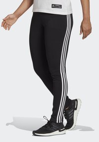Sorte leggings med tre vertikale hvite striper langs sidene, laget av stretchy stoff. Båret med svarte treningssko med hvite såler.